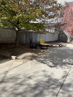 This is an outdoor walkway enviorment between the H wing building and the student center building. It is an enviorment made primarily of concrete and brick. there is a strip of natural space where trees are growing and underneath are some wooden chairs and benches. This space is used for leisure and comminity, the seating areas contain multiple chairs allowing for groups of people to gather but the location makes it so the enviorment is usually quite quiet.